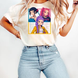 May include: Cream-colored t-shirt featuring a colorful anime-style graphic of three female characters. The graphic is divided into three sections with each character in a different pose. The word "VENOM" is visible on one of the character's shirt.
