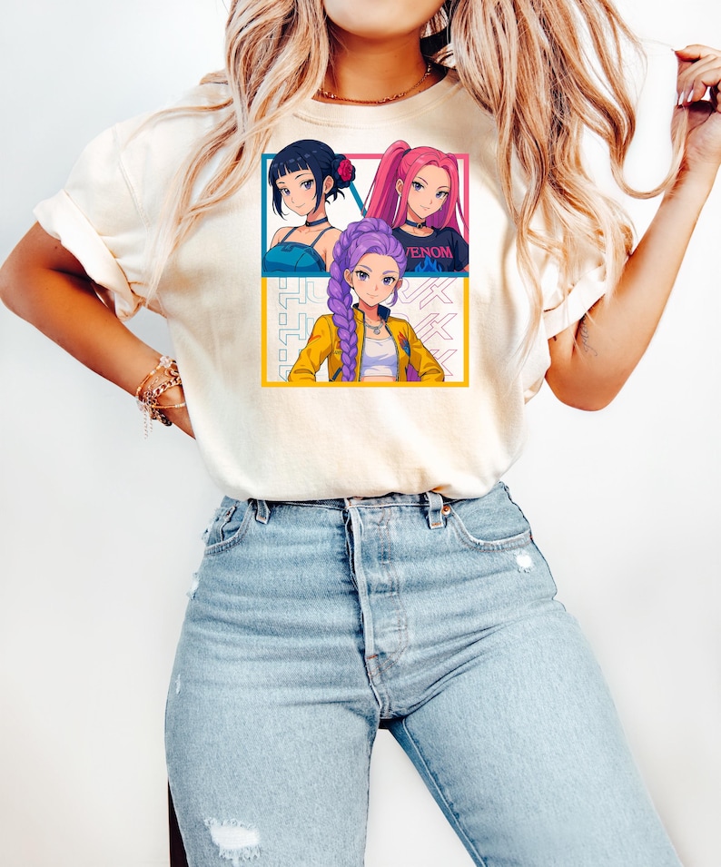 May include: Cream-colored t-shirt featuring a colorful anime-style graphic of three female characters. The graphic is divided into three sections with each character in a different pose. The word "VENOM" is visible on one of the character's shirt.