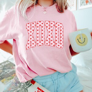 May include: A light pink sweatshirt with the letters "XOXO" in red, filled with tiny red hearts. The person is holding a white mug with a yellow smiley face. The person is wearing light blue shorts.