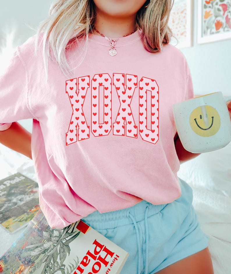 May include: A light pink sweatshirt with the letters "XOXO" in red, filled with tiny red hearts. The person is holding a white mug with a yellow smiley face. The person is wearing light blue shorts.