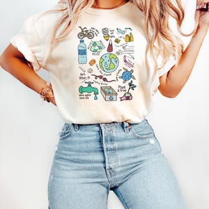 Pode incluir: Camiseta creme com um gr&aacute;fico colorido que promove a conscientiza&ccedil;&atilde;o ambiental. O design inclui ilustra&ccedil;&otilde;es de uma bicicleta, garrafa de &aacute;gua, globo, tartaruga e s&iacute;mbolos de reciclagem, juntamente com texto como "RECYCLE", "REDUCE" e "Plant a tree".