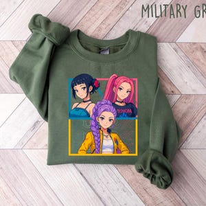 May include: Military green sweatshirt featuring a graphic of three anime-style girls with different hair colors. The graphic is framed in yellow and blue. The sweatshirt is made of a soft material.
