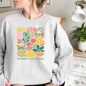 May include: A gray sweatshirt with a colorful floral design and the text "Community is Resistance".