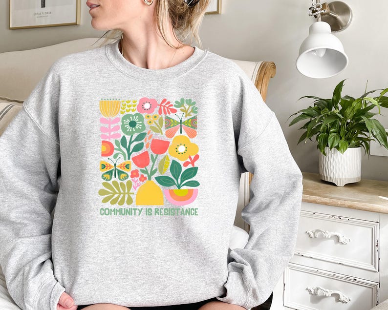 May include: A gray sweatshirt with a colorful floral design and the text "Community is Resistance".