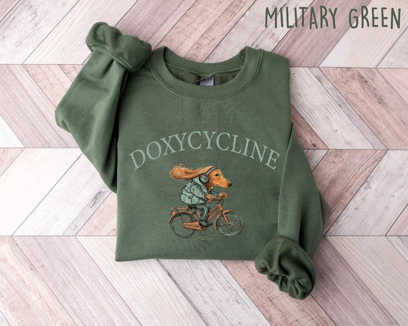 K&ouml;nnte beinhalten: Ein olivgr&uuml;nes Sweatshirt mit dem Aufdruck "DOXYCYCLINE" auf der Vorderseite. Ein Cartoon-Dackel mit Kopfh&ouml;rern f&auml;hrt Fahrrad.