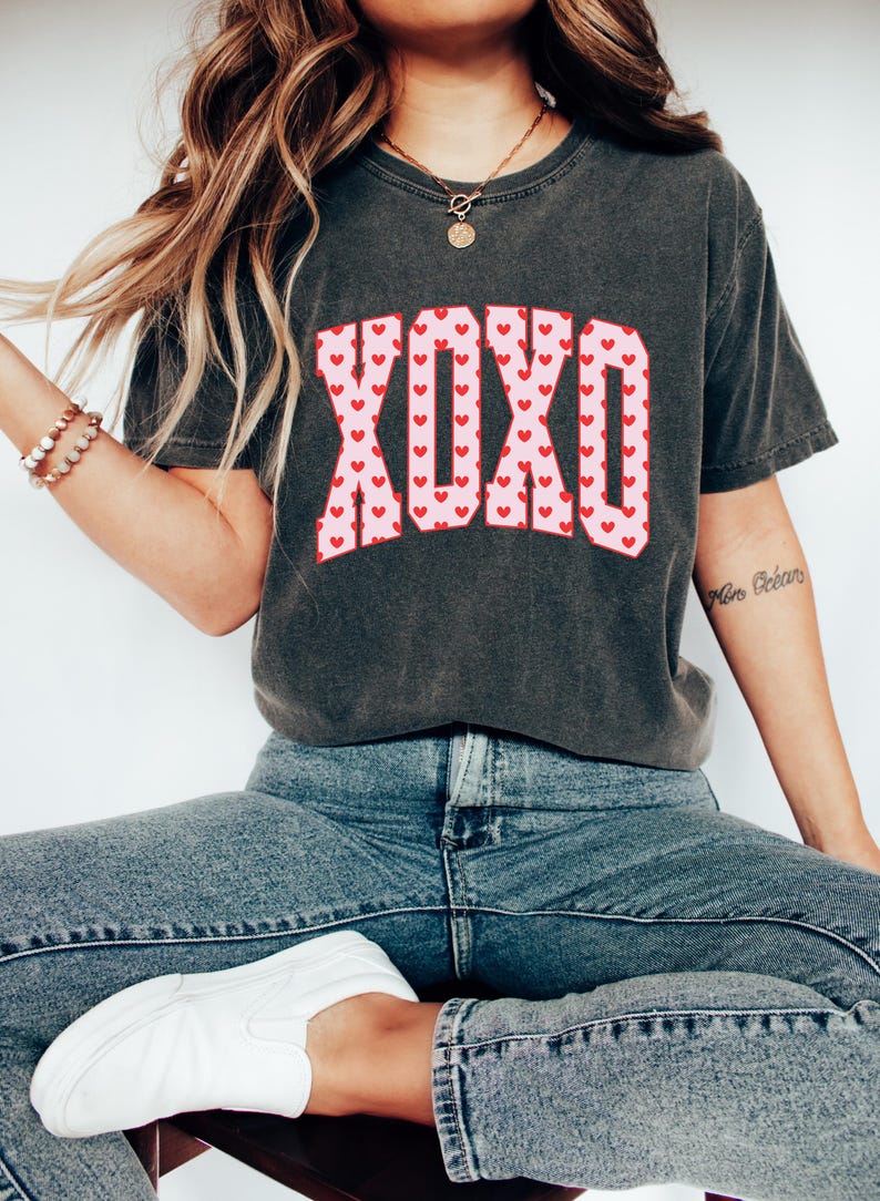 May include: Dark gray t-shirt with the word "XOXO" in a pink font filled with red hearts. The shirt is worn with blue jeans and white shoes. A gold necklace and bracelets are also visible.