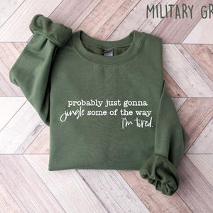 May include: Military green sweatshirt with the text "probably just gonna jingle some of the way I'm tired" in white script. The sweatshirt is folded on a light wood-grain surface.