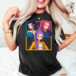May include: Black t-shirt featuring a colorful graphic of three anime-style women. The graphic is framed in blue and yellow, with the text "JUK" in the background. One woman has dark hair, another has pink hair, and the third has purple hair.