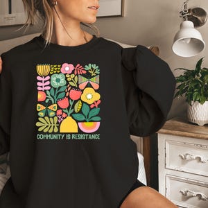 May include: Black sweatshirt with a colorful floral and butterfly design. The text "Community is Resistance" is printed below the design.