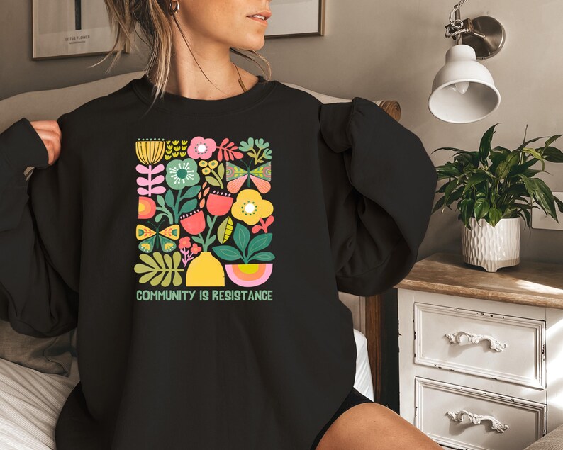 May include: Black sweatshirt with a colorful floral and butterfly design. The text "Community is Resistance" is printed below the design.