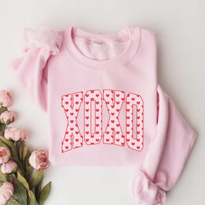 May include: A light pink sweatshirt with the letters "XOXO" in a red heart pattern. The sweatshirt is folded, with the sleeves visible. Pink flowers are arranged near the sweatshirt.