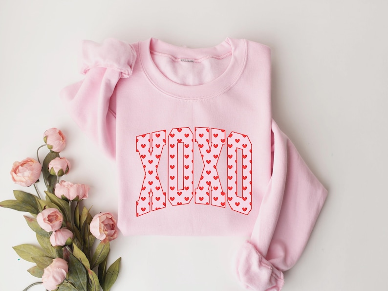May include: A light pink sweatshirt with the letters "XOXO" in a red heart pattern. The sweatshirt is folded, with the sleeves visible. Pink flowers are arranged near the sweatshirt.
