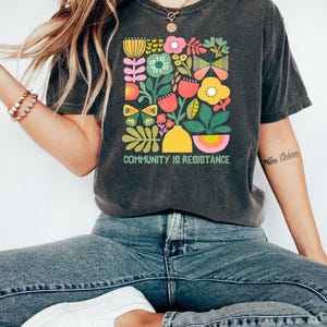 May include: A dark gray t-shirt with a colorful floral design and the text "COMMUNITY IS RESISTANCE".