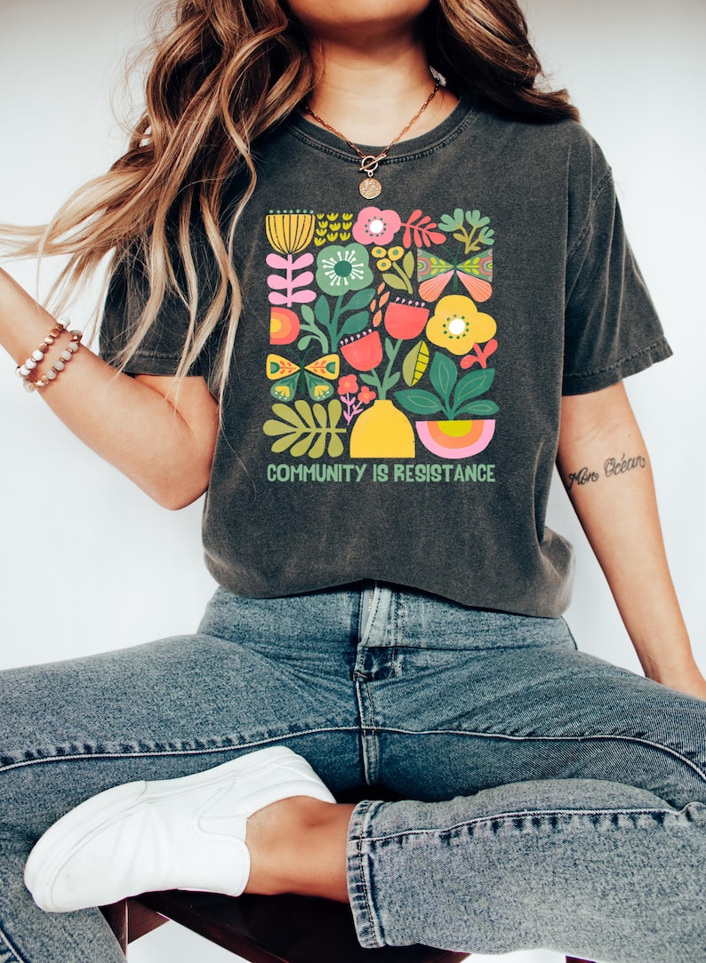 May include: A dark gray t-shirt with a colorful floral design and the text "COMMUNITY IS RESISTANCE".