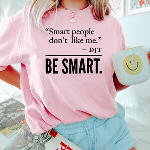Może przedstawiać: Jasnor&oacute;żowa bluza z nadrukiem "Smart people don't like me. - DJT BE SMART." w czarnym kolorze. Osoba trzyma biały kubek z ż&oacute;łtą uśmiechniętą buzią.