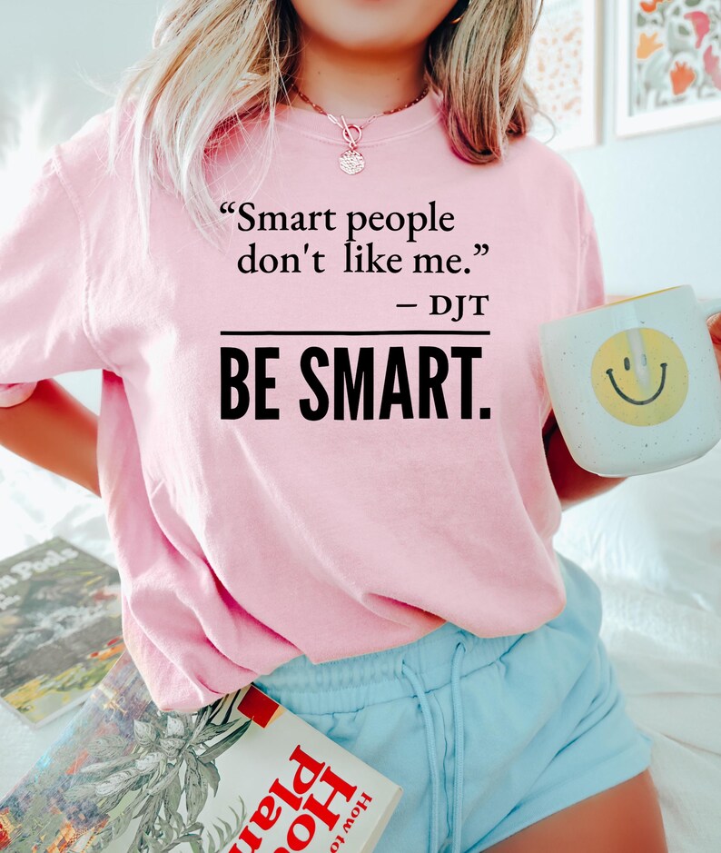 Może przedstawiać: Jasnor&oacute;żowa bluza z nadrukiem "Smart people don't like me. - DJT BE SMART." w czarnym kolorze. Osoba trzyma biały kubek z ż&oacute;łtą uśmiechniętą buzią.