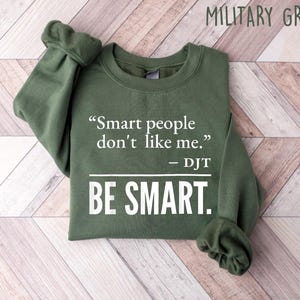 Może przedstawiać: Zielona wojskowa bluza z cytatem "Smart people don't like me." przypisanym do DJT i słowami "BE SMART." w kolorze białym. Bluza jest złożona na jasnej powierzchni z usłojeniem drewna.