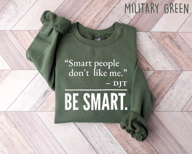 Może przedstawiać: Zielona wojskowa bluza z cytatem "Smart people don't like me." przypisanym do DJT i słowami "BE SMART." w kolorze białym. Bluza jest złożona na jasnej powierzchni z usłojeniem drewna.