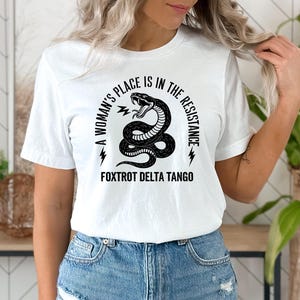 May include: White t-shirt with black text that reads "A woman's place is in the resistance" with a snake graphic and the words "Foxtrot Delta Tango" below.