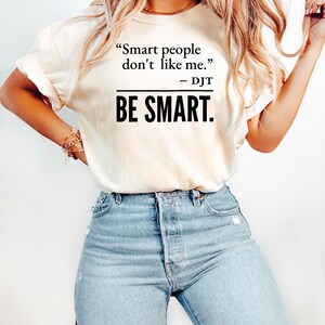Może przedstawiać: Kremowy t-shirt z cytatem "Smart people don't like me. - DJT" wydrukowanym na czarno. Poniżej słowa "BE SMART" są wydrukowane pogrubioną, czarną czcionką. Modelka ma na sobie jasnoniebieskie dżinsy.