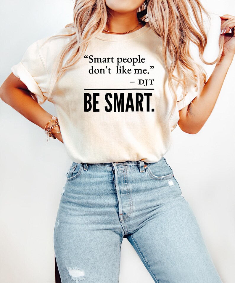 Może przedstawiać: Kremowy t-shirt z cytatem "Smart people don't like me. - DJT" wydrukowanym na czarno. Poniżej słowa "BE SMART" są wydrukowane pogrubioną, czarną czcionką. Modelka ma na sobie jasnoniebieskie dżinsy.