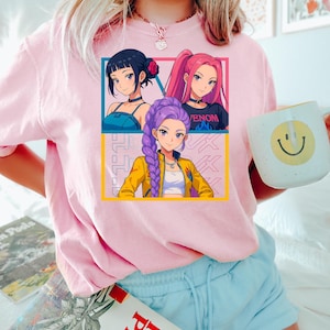 May include: Pink short-sleeved t-shirt featuring a graphic of three anime-style women with different hairstyles and outfits. The shirt also has Japanese text and a yellow and blue border. A white mug with a smiley face is held in the hand.