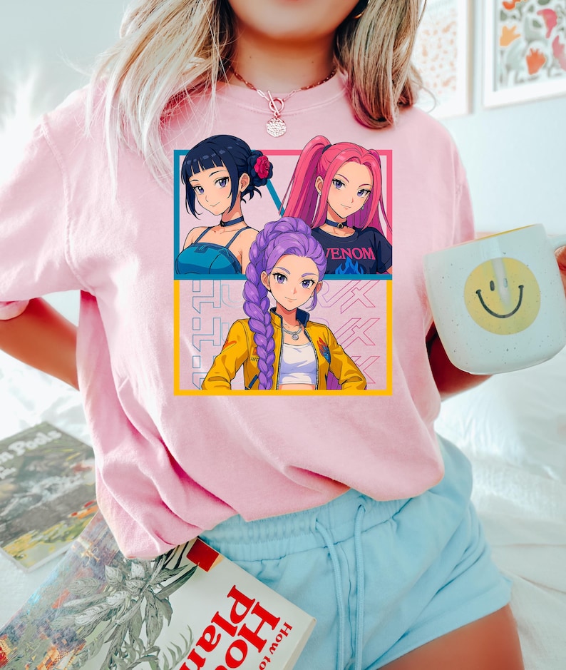 May include: Pink short-sleeved t-shirt featuring a graphic of three anime-style women with different hairstyles and outfits. The shirt also has Japanese text and a yellow and blue border. A white mug with a smiley face is held in the hand.