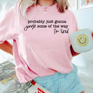 May include: A light pink t-shirt with the text "probably just gonna jingle some of the way, I'm tired." A white mug with a yellow smiley face is held in the hand. The person is wearing light blue shorts.