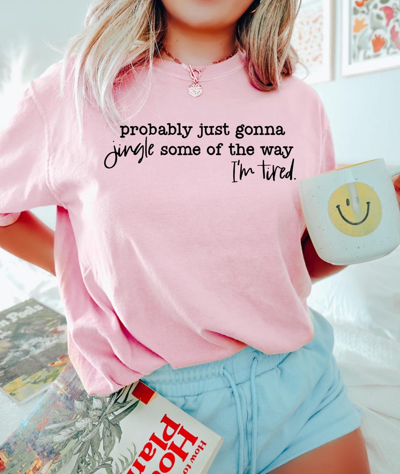 May include: A light pink t-shirt with the text "probably just gonna jingle some of the way, I'm tired." A white mug with a yellow smiley face is held in the hand. The person is wearing light blue shorts.