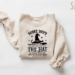 May include: Sand-colored sweatshirt with the text "SOME DAYS YOU JUST HAVE TO PUT ON THE HAT to remind them who THEY'RE DEALING WITH" in black. The graphic includes a witch hat and broom. A cozy, casual top.