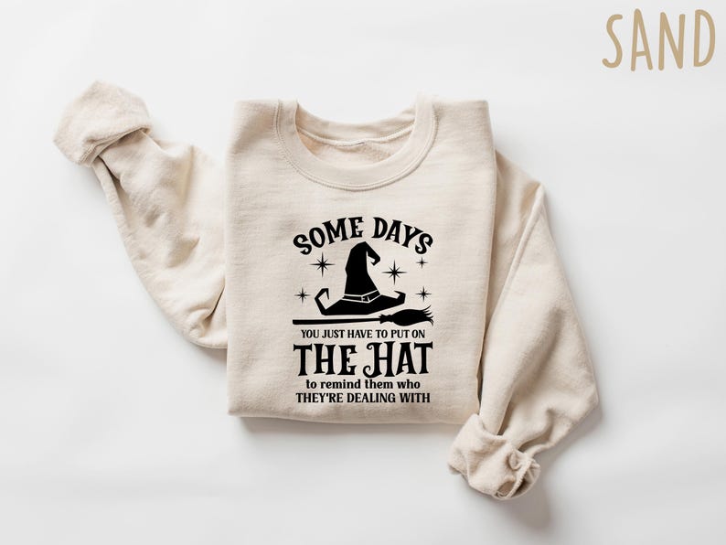 May include: Sand-colored sweatshirt with the text "SOME DAYS YOU JUST HAVE TO PUT ON THE HAT to remind them who THEY'RE DEALING WITH" in black. The graphic includes a witch hat and broom. A cozy, casual top.