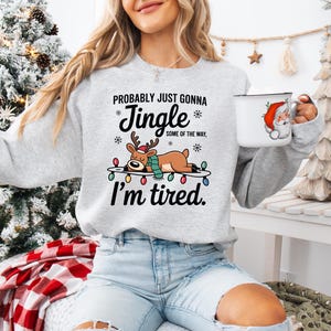 May include: A light gray sweatshirt with the text "Probably Just Gonna Jingle Some of the Way, I'm Tired" and a graphic of a reindeer lying on a string of Christmas lights. A white mug with a Santa Claus graphic is held in the hand.