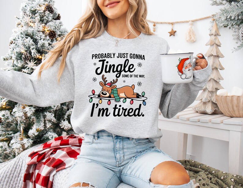 May include: A light gray sweatshirt with the text "Probably Just Gonna Jingle Some of the Way, I'm Tired" and a graphic of a reindeer lying on a string of Christmas lights. A white mug with a Santa Claus graphic is held in the hand.