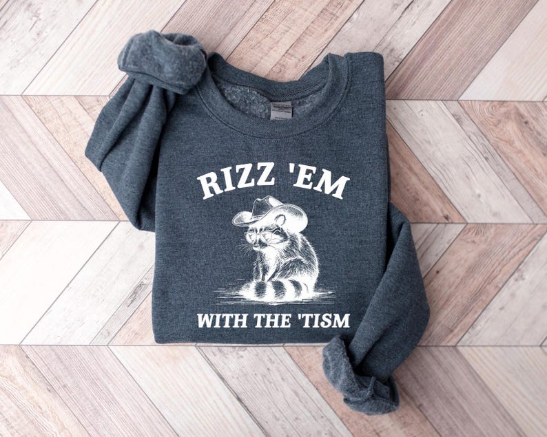 K&ouml;nnte beinhalten: Ein dunkelgrauer Pullover mit dem Text "Rizz 'Em With The 'Tism" und einem Cartoon-Waschb&auml;ren, der einen Cowboyhut tr&auml;gt.