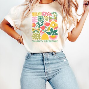 May include: A woman wearing a cream colored t-shirt with a colorful floral design and the text "Community is Resistance". She is also wearing light blue jeans.