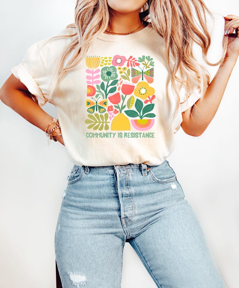 May include: A woman wearing a cream colored t-shirt with a colorful floral design and the text "Community is Resistance". She is also wearing light blue jeans.