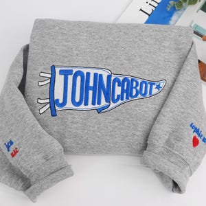 Puede incluir: Una sudadera gris con un bander&iacute;n azul y blanco que dice "JOHN CABOT". La manga tiene el texto "sophie m." y un coraz&oacute;n rojo. La otra manga tiene el texto "jca mac."