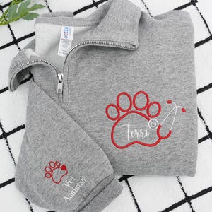 May include: A gray quarter-zip sweatshirt with a red paw print design and the name "Terri" embroidered on the front. The sleeve features a smaller paw print and the words "Vet Assistant".