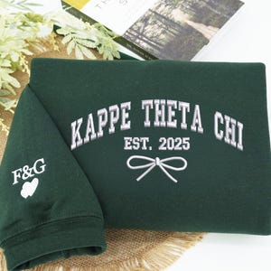 May include: A dark green sweatshirt with the text "KAPPE THETA CHI EST. 2025" embroidered in silver on the front. A small bow is embroidered below the text. The left sleeve has the letters "F&G" and a heart embroidered in white.