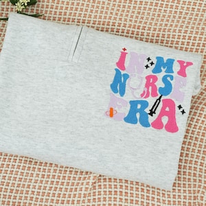 May include: A folded light gray sweatshirt with the embroidered text "I <3 MY NURSE ERA" in pink, blue, and lavender. The design includes a syringe, a stethoscope, and a cross.