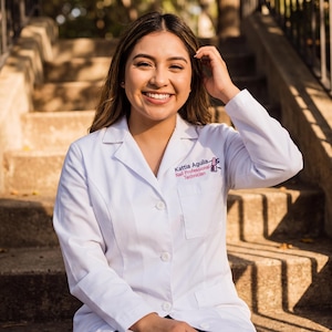 May include: A smiling person wearing a white lab coat with the text "Kattia Aguila Nail Professional Technician" in pink. The person is sitting on concrete steps outdoors.