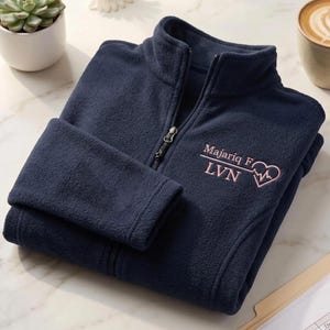 May include: A navy blue fleece jacket with a high collar and a full-length front zip. The chest features embroidered light pink text that reads Majariq F LVN, accompanied by a line graphic ending in a heart shape with an EKG pulse line inside.