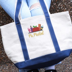 Canvas tote bag with dark blue handles, embroidered apple, crayons, pencil, ruler, and paintbrush, personalized with name “Ms. Medal,” displayed outdoors.