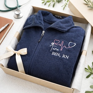 Könnte beinhalten: Marineblaue Fleecejacke mit durchgehendem Reißverschluss, präsentiert in einer Geschenkbox mit cremefarbenem Band. Die Jacke hat ein gesticktes Stethoskop-Design mit dem Namen "Tata" und den Abschlüssen "BSN, RN". Ein Stethoskop und ein Notizblock sind im Hintergrund zu sehen.