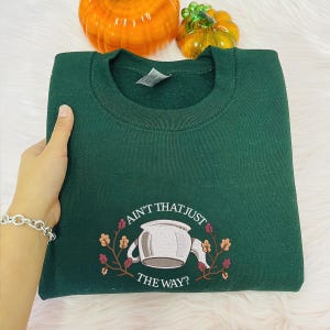 May include: A dark green sweatshirt with the text "Ain't that just the way?" and a white teapot graphic with brown leaves and flowers.