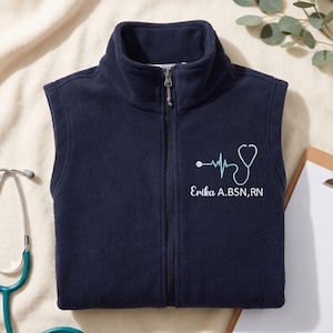 Può includere: Gilet in pile blu navy con cerniera intera e collo alto. Presenta un design a stetoscopio e battito cardiaco azzurro, con il nome "Erika A. BSN, RN" ricamato sotto. Sullo sfondo è visibile uno stetoscopio turchese.