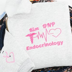 May include: A light gray sweatshirt with pink embroidered text and graphics. The text reads "Kim DNP Endocrinology" with a medical symbol and a heartbeat line ending in a heart. A smaller section of the sweatshirt shows a stethoscope graphic.