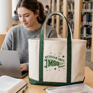 May include: Cream-colored canvas tote bag with green accents and handles. The bag features the text "Michigan Univ. MSU Harper" in green. The bag is sitting on a wooden table next to a laptop and an open book.