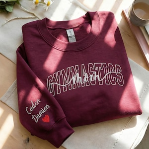 May include: A maroon sweatshirt with the word "GYMNASTICS" in white block letters and "Mom" in cursive. The left sleeve has the names "Caden" and "Damien" embroidered in white, with a red heart below. The sweatshirt is folded on a light-colored surface.