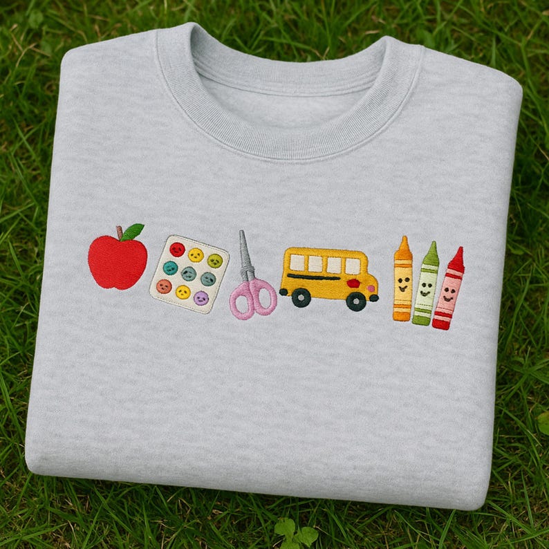 Folded grey sweatshirt with colorful embroidered school icons including apple, paint palette, scissors, school bus, and crayons – perfect back-to-school gift for teachers.